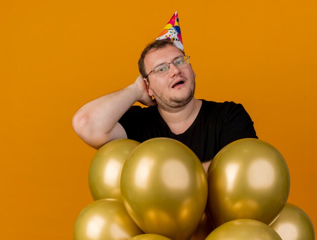 Free photo surprised adult slavic man in optical glasses wearing birthday cap puts hand on head behind and stands with helium balloons