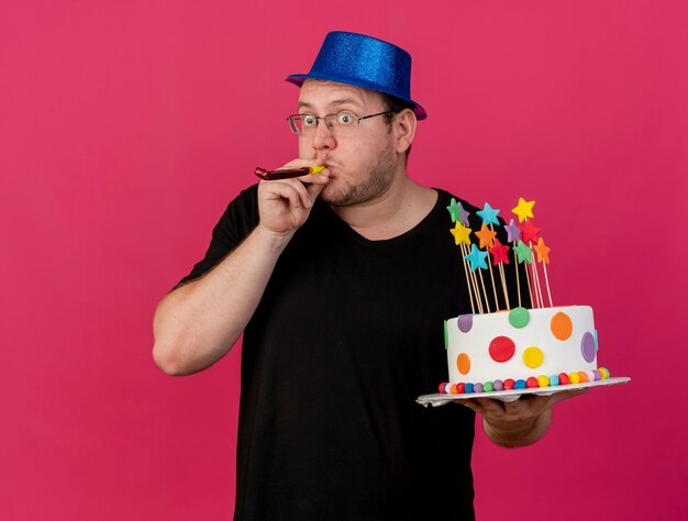 Surprised adult slavic man in optical glasses wearing blue party hat holds birthday cake blowing party whistle 