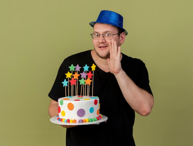 Surprised adult slavic man in optical glasses wearing blue party hat keeps hand close to mouth and holds birthday cake looking at camera 