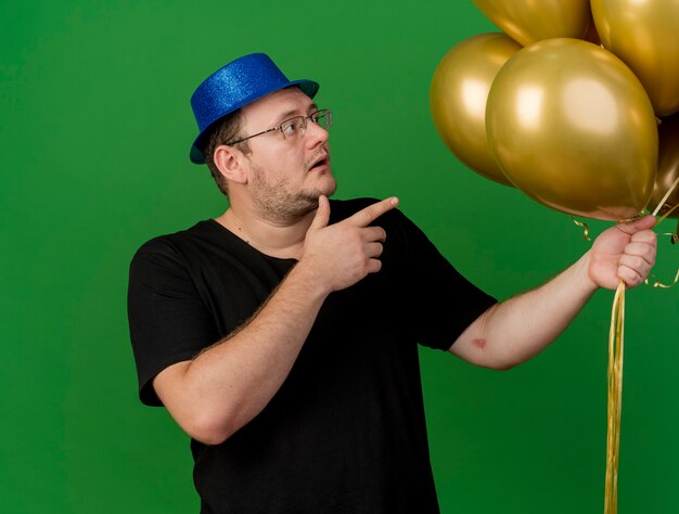 Surprised adult slavic man in optical glasses wearing blue party hat looks and points at helium balloons 