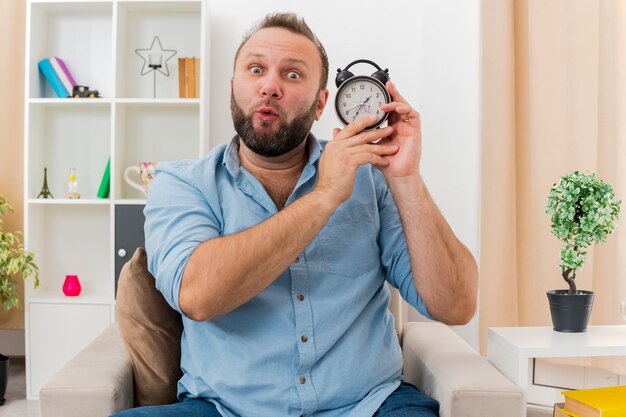 Surprised adult slavic man sits on armchair holding alarm clock looking at camera inside the living room