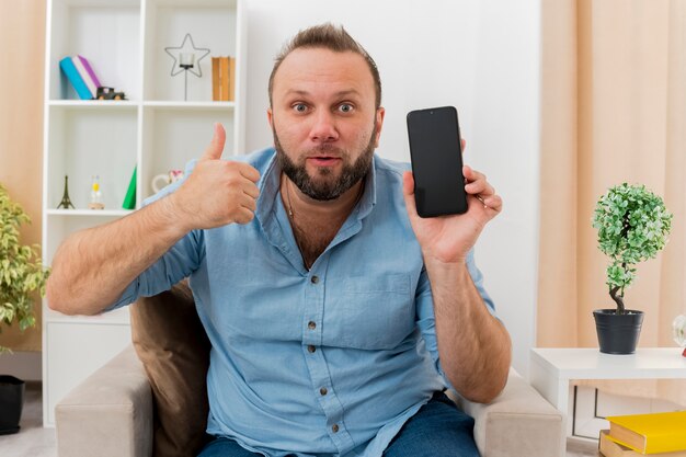 Surprised adult slavic man sits on armchair holding phone and thumbs up inside the living room