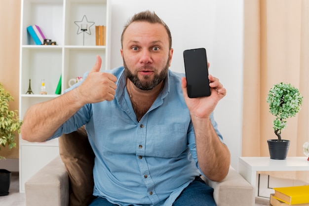 Удивленный взрослый славянский мужчина сидит на кресле с телефоном и показывает палец вверх в гостиной