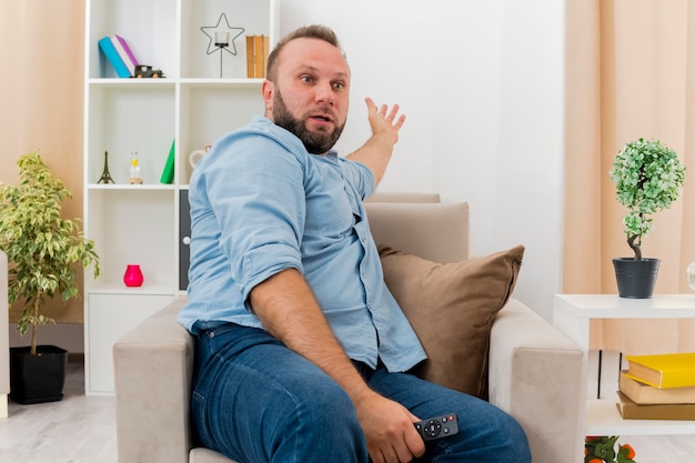 Surprised adult slavic man sits on armchair holding tv remote and pointing behind inside living room