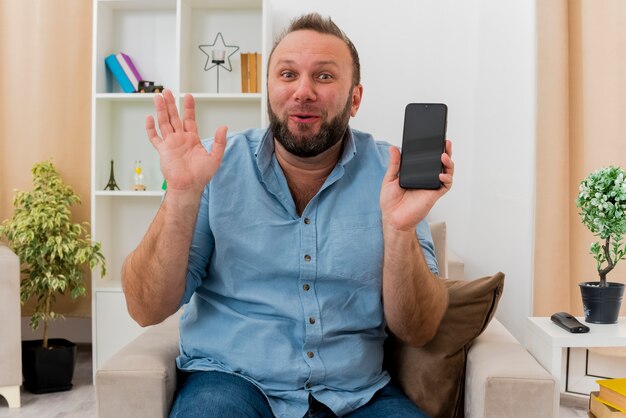 Surprised adult slavic man sits on armchair raising hand and holding phone inside the living room