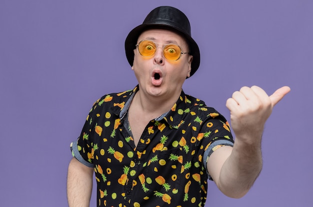 Surprised adult slavic man with black top hat wearing sunglasses pointing at side