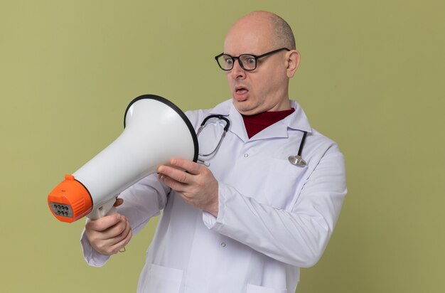 Free photo surprised adult slavic man with optical glasses in doctor uniform with stethoscope holding and looking at loud speaker