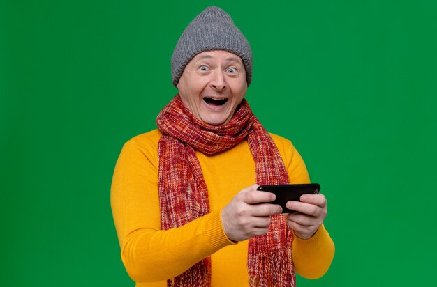 Surprised adult slavic man with winter hat and scarf around his neck holding phone 