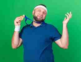 Foto gratuita sorpreso adulto slavo sportivo uomo che indossa fascia e braccialetti che tengono medaglia d'oro isolata sulla parete verde con spazio copia