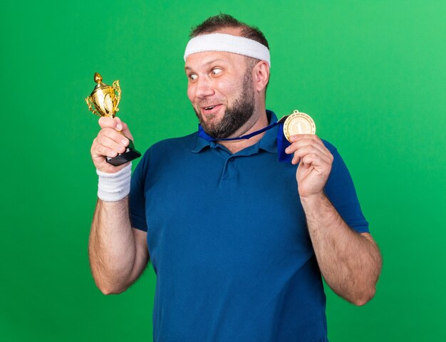 surprised adult slavic sporty man wearing headband and wristbands holding golden medal and looking at winner cup isolated on green wall with copy space