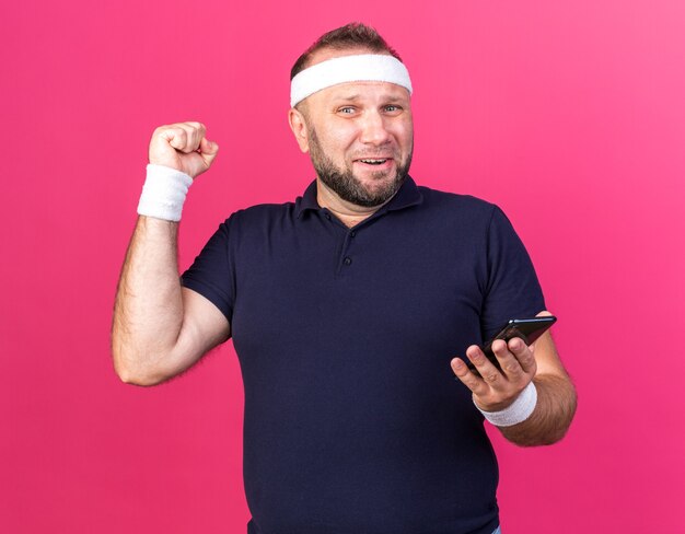 surprised adult slavic sporty man wearing headband and wristbands holding phone and keeping fist up isolated on pink wall with copy space