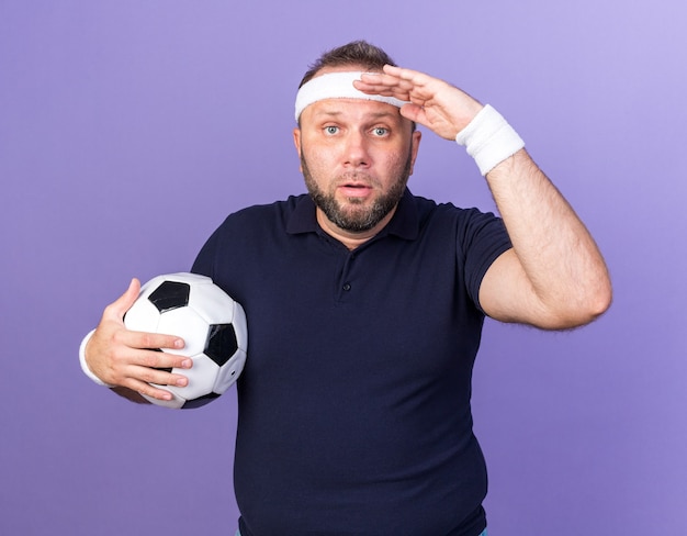 удивленный взрослый славянский спортивный мужчина с повязкой на голову и браслетами, держащий ладонь у лба и держащий мяч, изолированный на фиолетовой стене с копией пространства