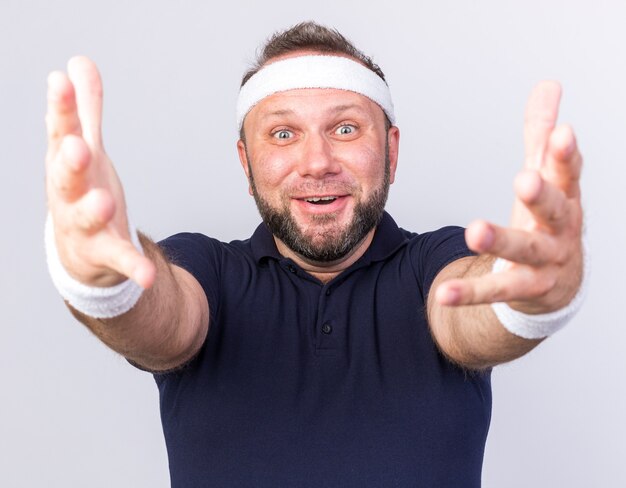 surprised adult slavic sporty man wearing headband and wristbands stretching out hands isolated on white wall with copy space