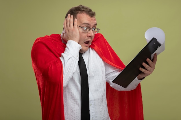 Surprised adult slavic superhero man in red cape wearing glasses and tie holding and looking at clipboard putting hand on head isolated on olive green background