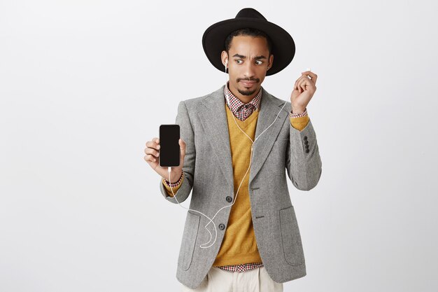 Surprised african-american guy take-off headphone confused and awkward, looking at smartphone screen, showing something strange on mobile display