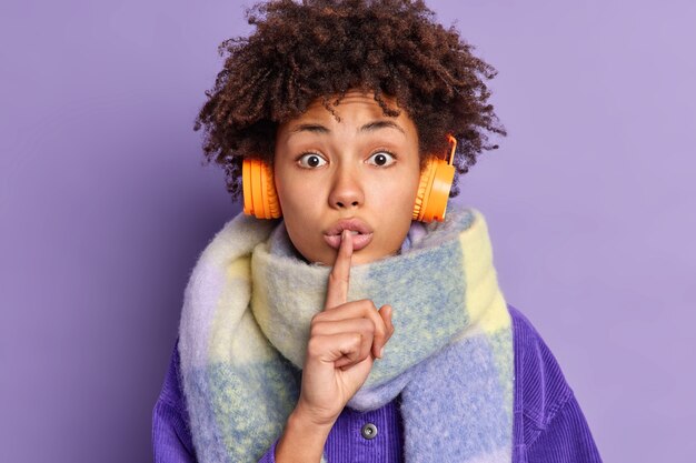 surprised African American woman presses index finger to lips makes silence gesture tells secret wears warm scarf around neck listens music via wireless headphones.