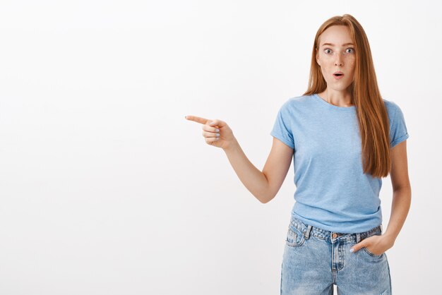 Surprised and amazed attractive ginger girl with freckles in blue t-shirt gasping with folded lips popping eyes and staring impressed pointing left thrilled