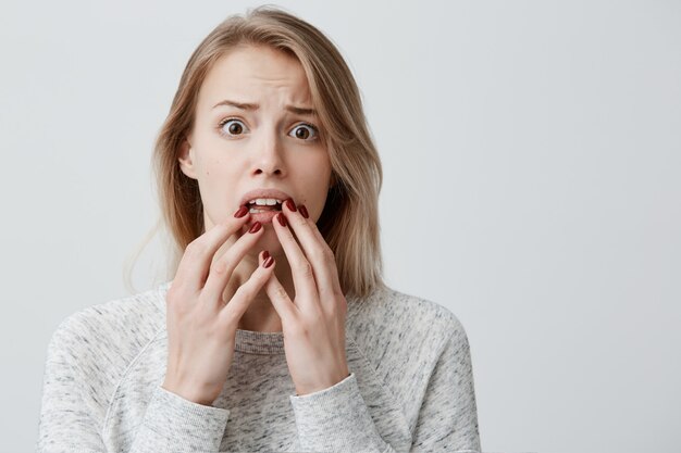 Surprised amazed attractive young blonde with dyed hair female wearingsweater having astonished face expression, covering her open mouth with hands