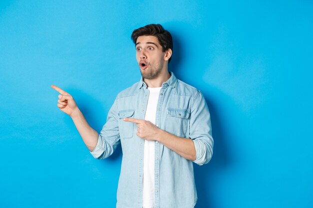 Surprised and amazed man looking at promotion, pointing fingers left at advertisement, standing over blue background.
