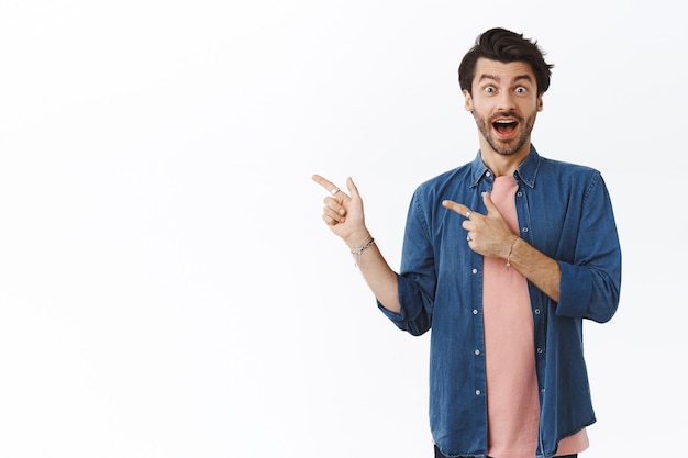 Surprised, amused good-looking man with beard, open mouth fascinated, raise eyebrows wondered, enjoy playing in amusement park, pointing fingers left, astonished with amazing promo, white wall