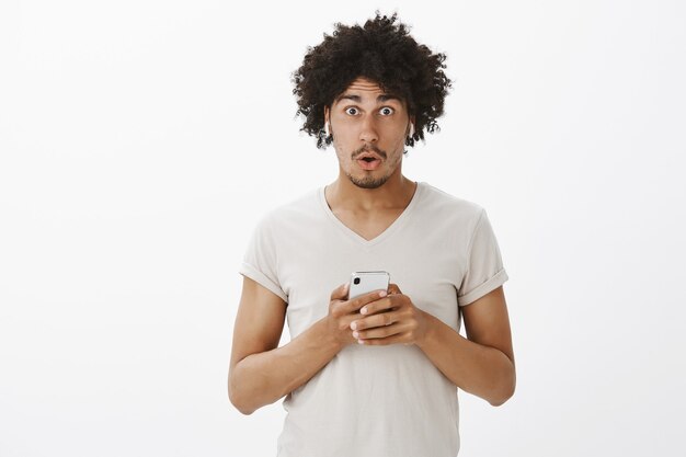 Surprised and amused handsome man in wireless headphones, hold smartphone, looking impressed