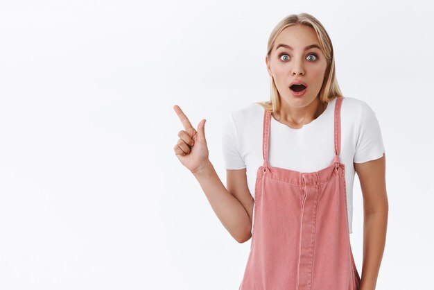 Surprised and amused young girl with blond hair pointing upper left corner fascinated with amazing cool new promo open mouth wondered and astonished look camera as telling unbelievable news