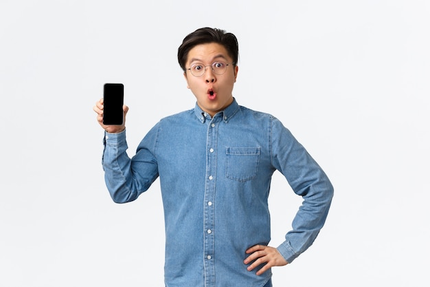 안경과 평상복 차림의 놀랍고 놀란 아시아 남성, 휴대폰 화면을 보여주고 와우, 새로운 스마트폰 응용 프로그램, 온라인 최신 뉴스, 흰색 배경에 대해 토론