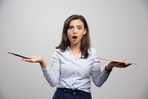 Surprised artist holding paintbrush and palette over gray wall. 