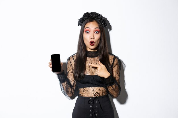 Surprised asian girl in black gothic dress with wreath, gasping amused and pointing finger at mobile phone display, showing halloween banner or promo, standing over white background.