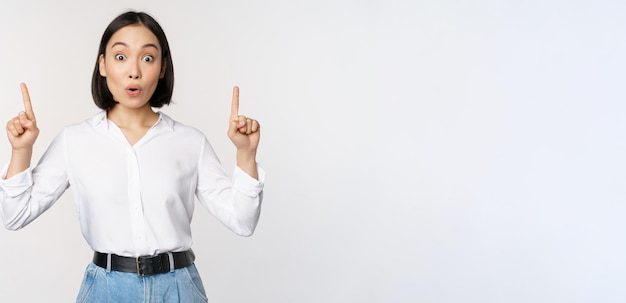 Surprised asian girl pointing fingers up showing top banner information or advertisement standing over white background in blouse Copy space