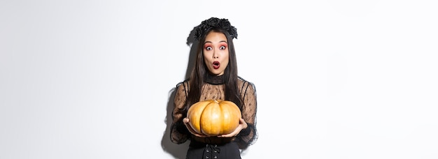 Surprised asian girl in witch costume holding pumpkin and gasping amazed at camera preparing for hal