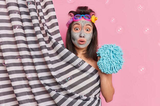 놀란 된 아시아 여자 얼굴 클레이 마스크 적용 날짜에 대 한 스펀지를 보유 하 고 샤워는 완벽 한 몸이 비누 거품과 분홍색 배경 위에 절연 휴식과 구호를 느낀다.