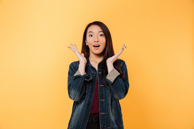 Surprised asian woman in denim jacket holding arms near face and looking at the camera over yellow background