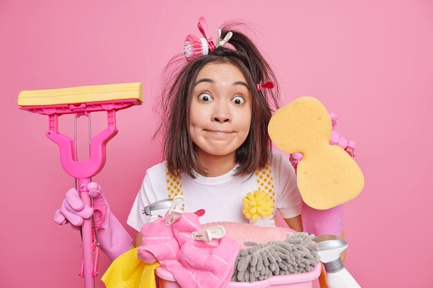 Surprised Asian woman holds sponge uses cleaning supplies poses with mop busy doing housework in new home poses against pink background. Domestic chores washing time and housekeeping concept