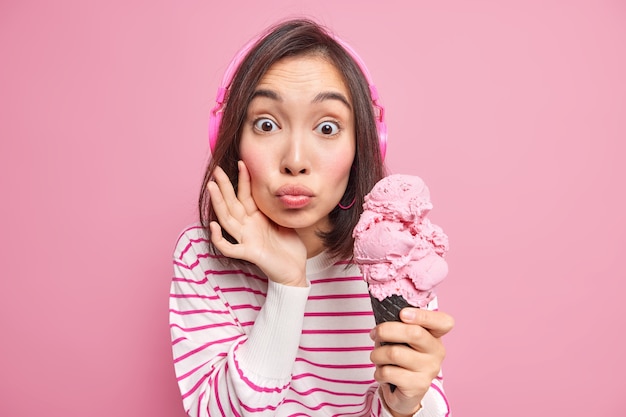 놀란 된 아시아 여자 입술 둥근 유지 맛있는 아이스크림 먹고 싶어 차가운 냉동 디저트 귀에 헤드폰을 착용 핑크 벽 위에 절연 캐주얼 스트라이프 점퍼. 여름 시간.
