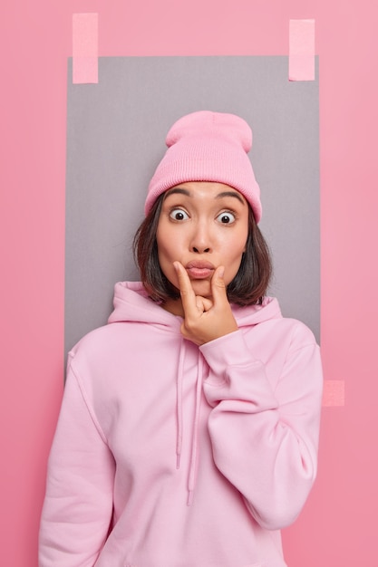 Surprised Asian woman makes grimace keeps lips rounded has bugged eyes wears hat and hoodie feels shocked poses against pink studio wall with stuck grey sheet of paper behind