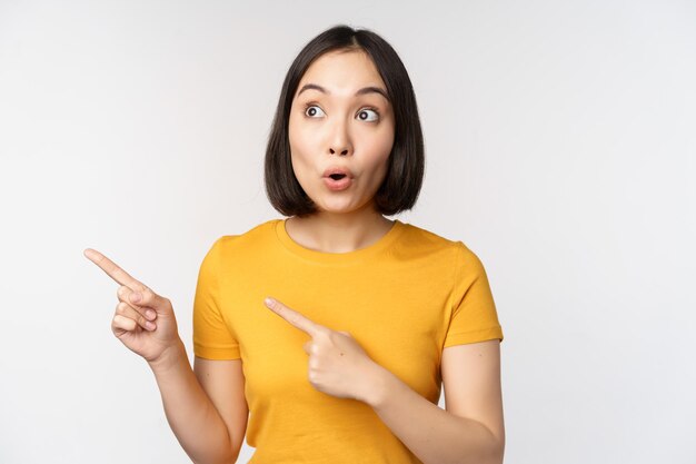 Surprised asian woman pointing and looking left at copy space promo sale showing advertisement with impressed face expression standing over white backround