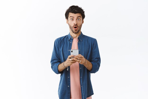 Surprised and astonished guy receives new smartphone, folding lip in wow and stare excited and shocked camera, see something amazing online, order it now, white wall