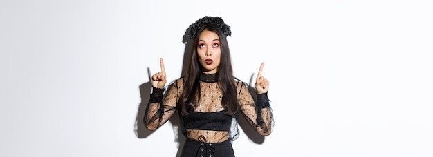 Удивленная привлекательная азиатка в костюме ведьмы, указывающая пальцем вверх, показывая промо баннера хэллоуина