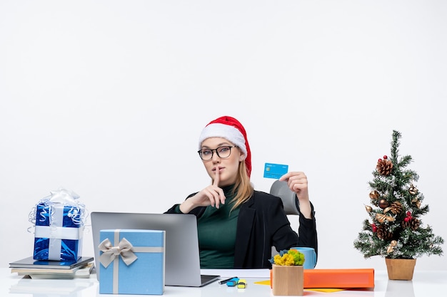 サンタクロースの帽子をかぶって、テーブルのクリスマスプレゼントに座って、オフィスで沈黙のジェスチャーを作る銀行カードを保持している眼鏡をかけて驚いた魅力的な女性