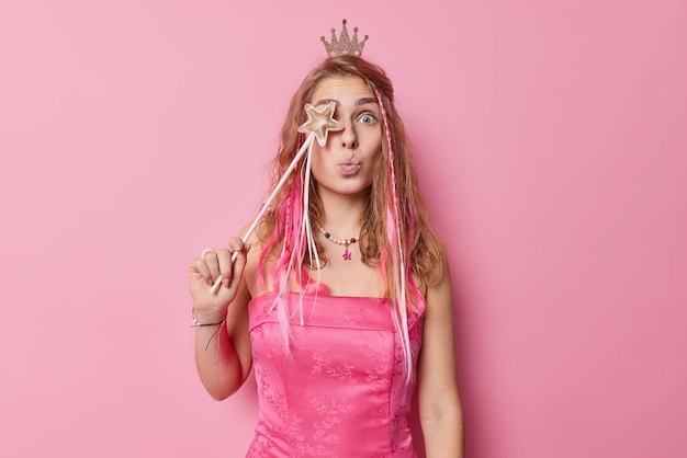 Surprised attractive young woman covers eye with magic wand wears crown dress and necklace pretends being magician or queen poses against pink bacjground. Party time and celebration concept.