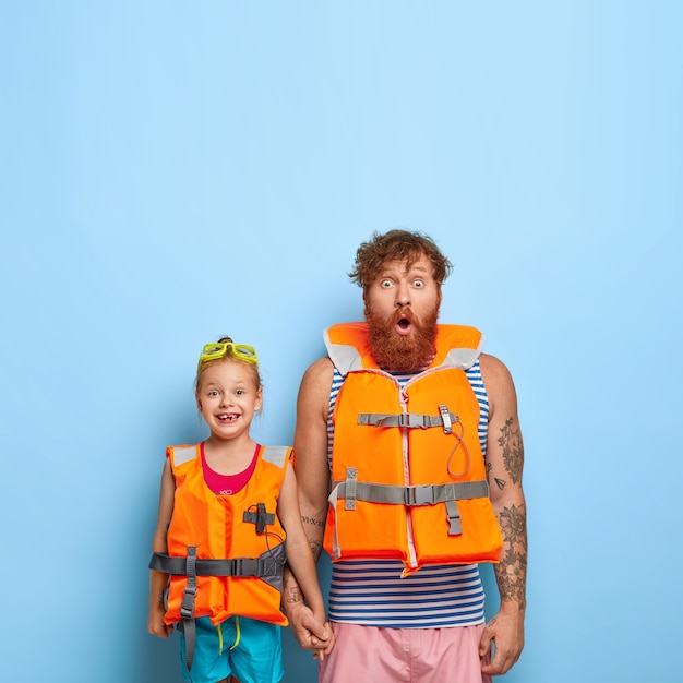 Surprised bearded dad with ginger beard, holds hand of little girl, wear protective lifejackets, ready for sea voyage, enjoy summer rest, stand against blue backgrond with copy space upwards