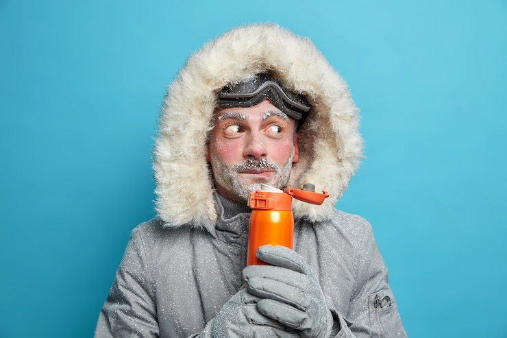 Free Photo surprised bearded european man covered with ice drinks hot beverage holds thermos wears snowboarding goggles and thermo jacket.