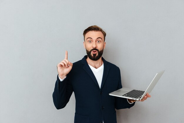 Удивленный бородатый человек в деловой одежде, держа ноутбук и имея идею, глядя на камеру над серым
