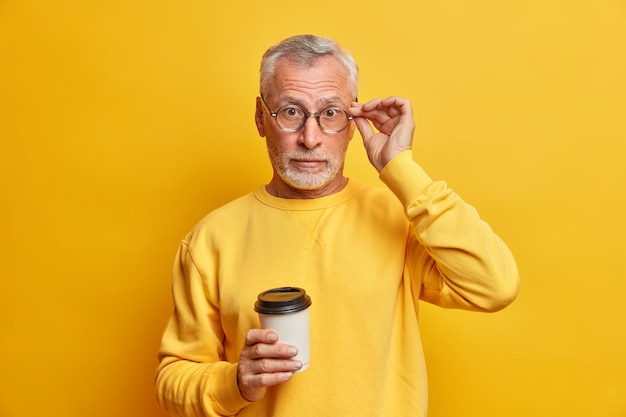 surprised bearded mature man keeps hand on spectacles drinks takeaway coffee hears astonishing news wears casual jumper isolated over vivid yellow wall