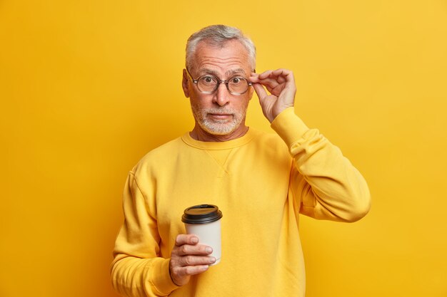驚いたひげを生やした成熟した男は、眼鏡を飲み続けますテイクアウトコーヒーは驚くべきニュースを聞きます鮮やかな黄色の壁の上に隔離されたカジュアルなジャンパーを着ています