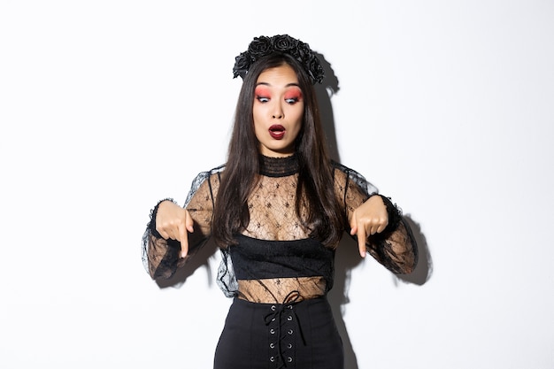 Surprised beatiful asian woman in black gothic dress and wreath, pointing fingers down and looking startled at something about halloween or trick or treat, standing over white background.