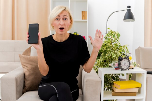 Surprised beautiful blonde russian woman sits on armchair raising hand and holding phone inside living room