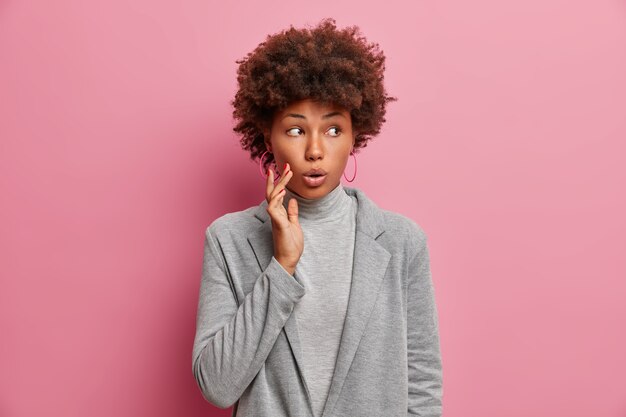 Surprised beautiful curly haired woman with wondered expression, gasps and looks away, being impressed and speechless, wears grey jacket