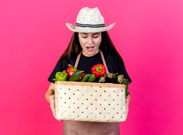 Бесплатное фото Удивленная красивая девушка-садовник в униформе в садовой шляпе держит и смотрит на корзину с овощами, изолированную на розовом